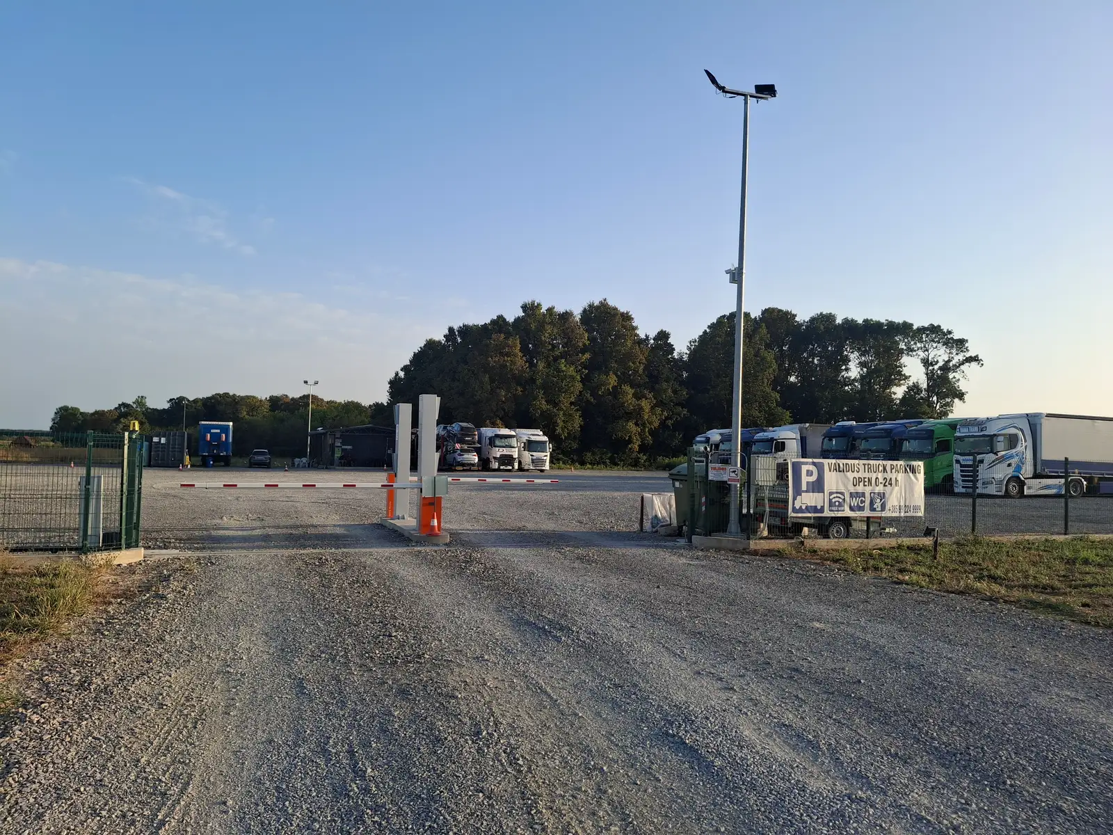 Ulaz u Validus Truck Parking: rampa, rasvjetni stup s kamerom i natpis „Validus Truck Parking – Open 0-24 h”, u pozadini parkirani kamioni